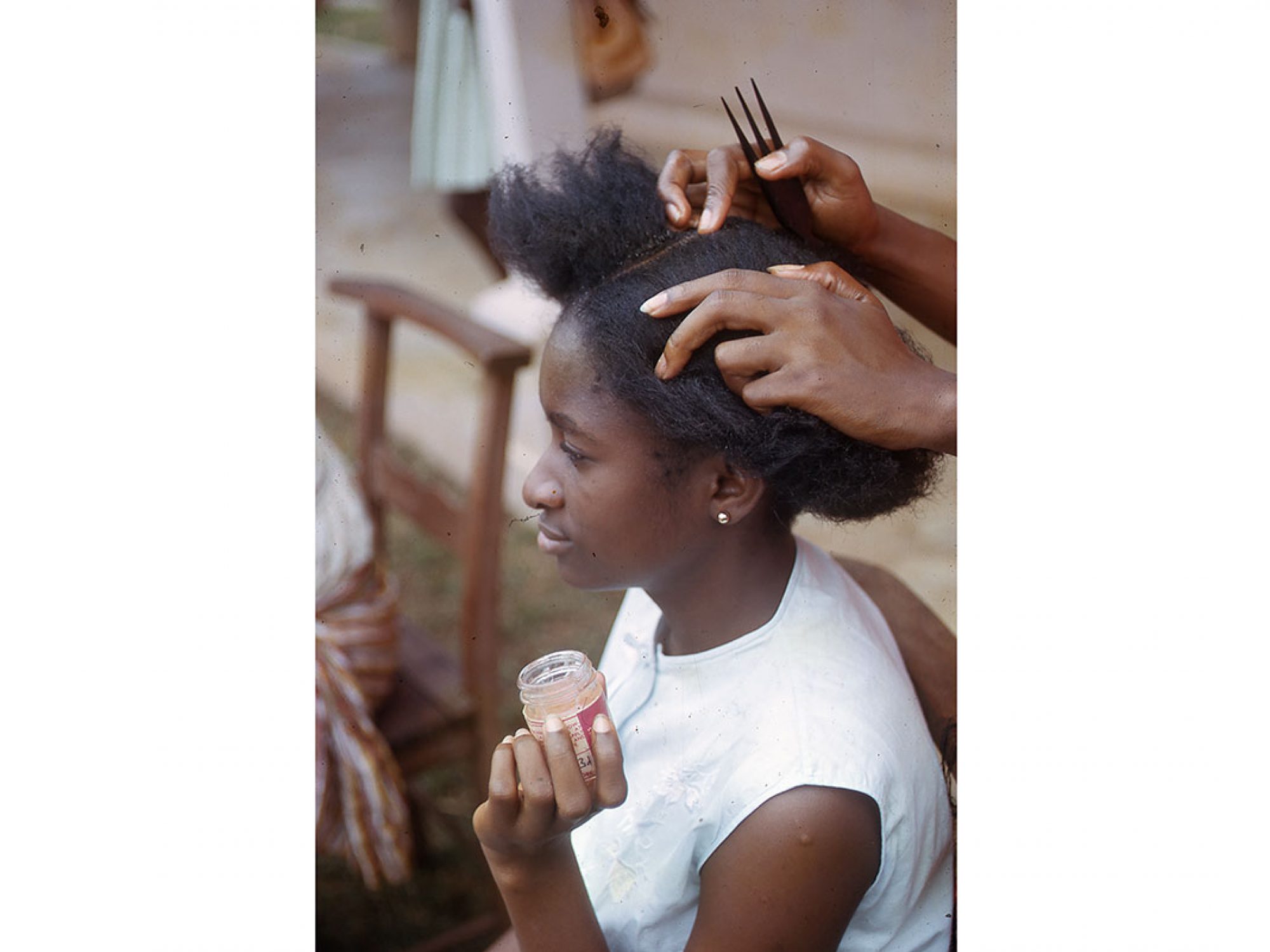 A woman is having her hair done - it has been parted by some hands (person unseen) who is also holding an afro comb. The girl wears a white top and is holding up a pot of hair product for the person doing her hair to use.