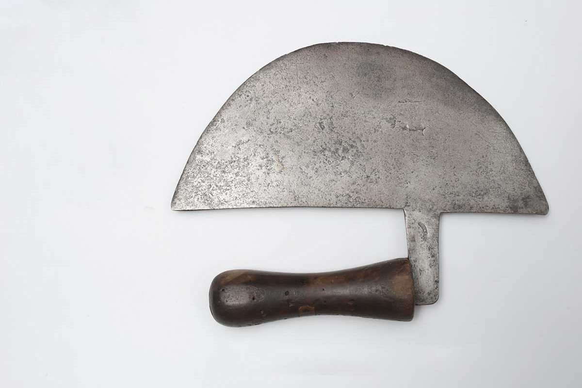 Cheese cutter with a thin crescent shaped iron blade and a short waisted wooden handle with a rounded end.