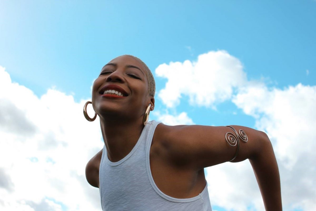 Demae in a white vest and hoop earrings looks happy against a blue sky