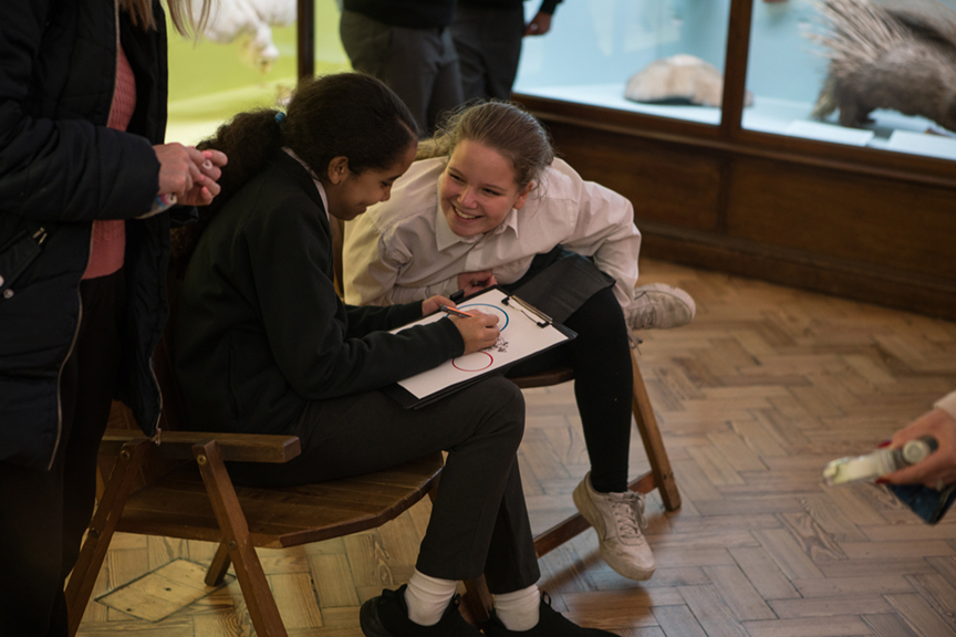 A child leans over to talk to her friend who is writing on a clipboard