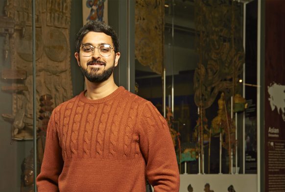 A man wearing an orange jumper and round glasses stands in a gallery, in front of a display case. He has dark hair and facial hair