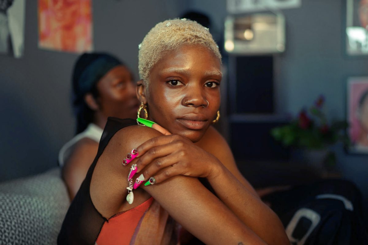 A black woman with cropped blonde hair looks at the camera as she holds her own arms. She has gold hoop earrings in and fake pink and green nails with are decorated with jewels