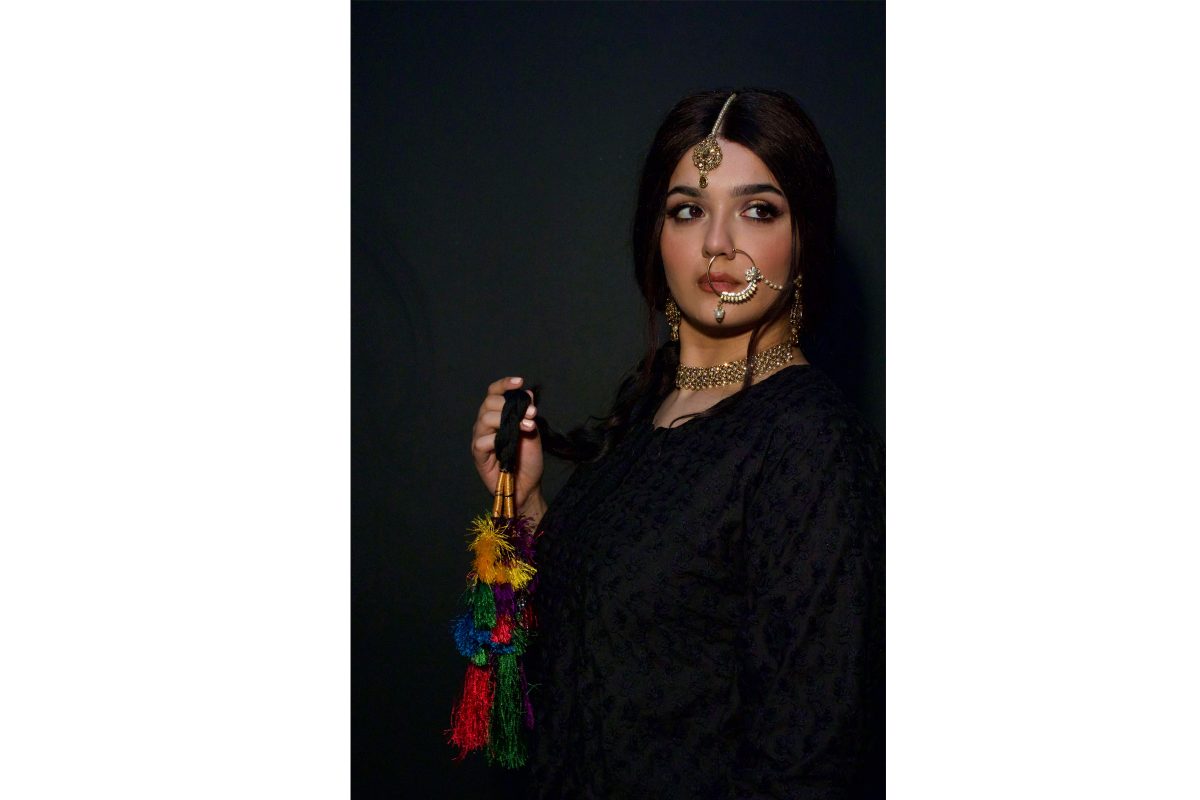 Portrait of a South Asian woman with long dark hair which ends in a colourful plait. She has gold jewellery on including a large hoop through her nose.