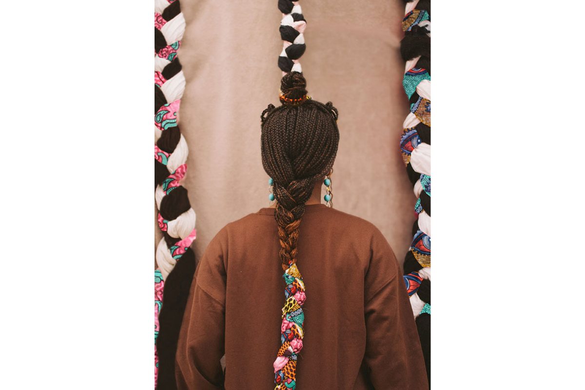 The back of a Black woman's head, her braids are plaited into fabric, and large pieces of plastic fabric are facing her