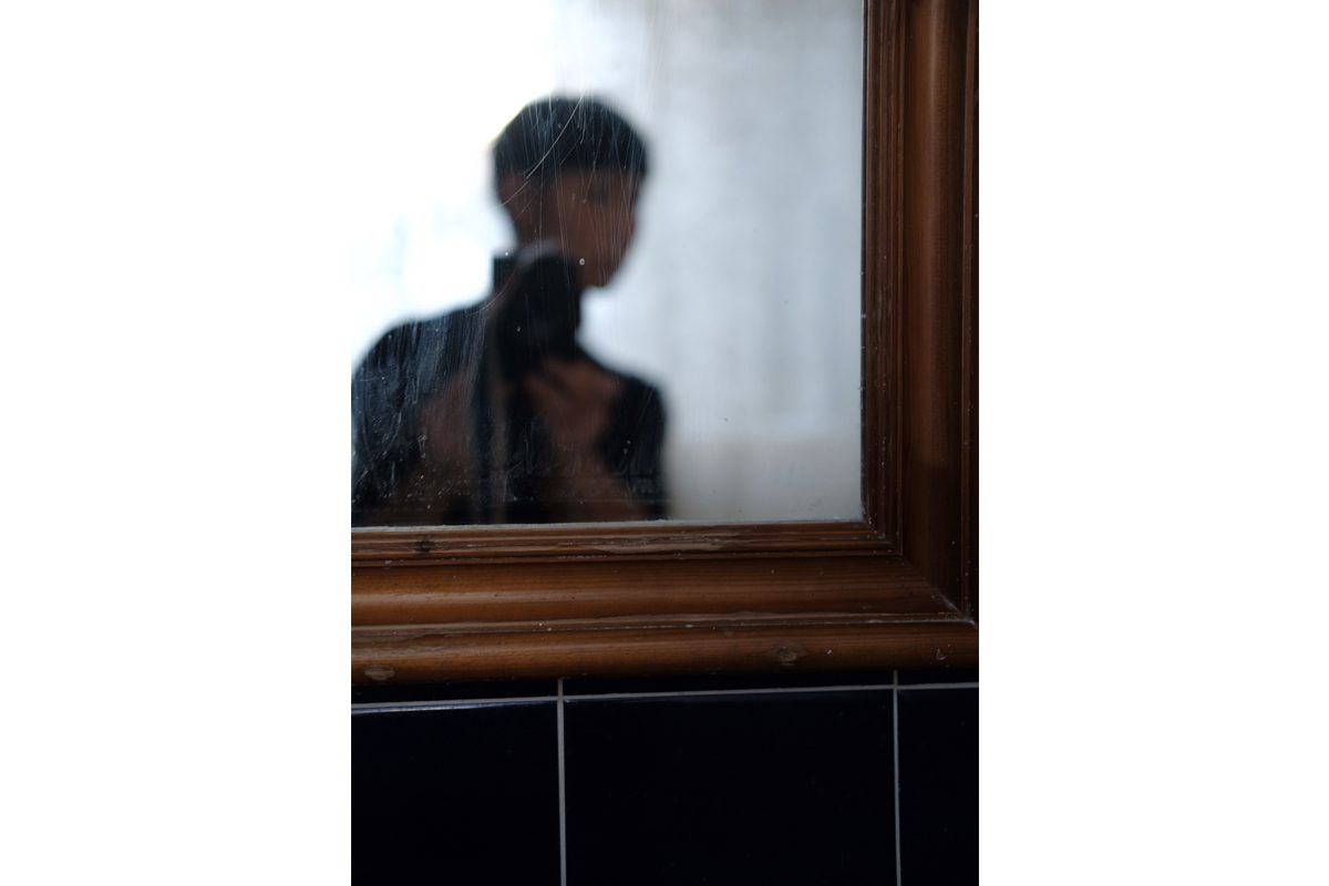 A woman takes a photo of herself in a mirror with a camera, wall is in focus but the image in the mirror is out of focus