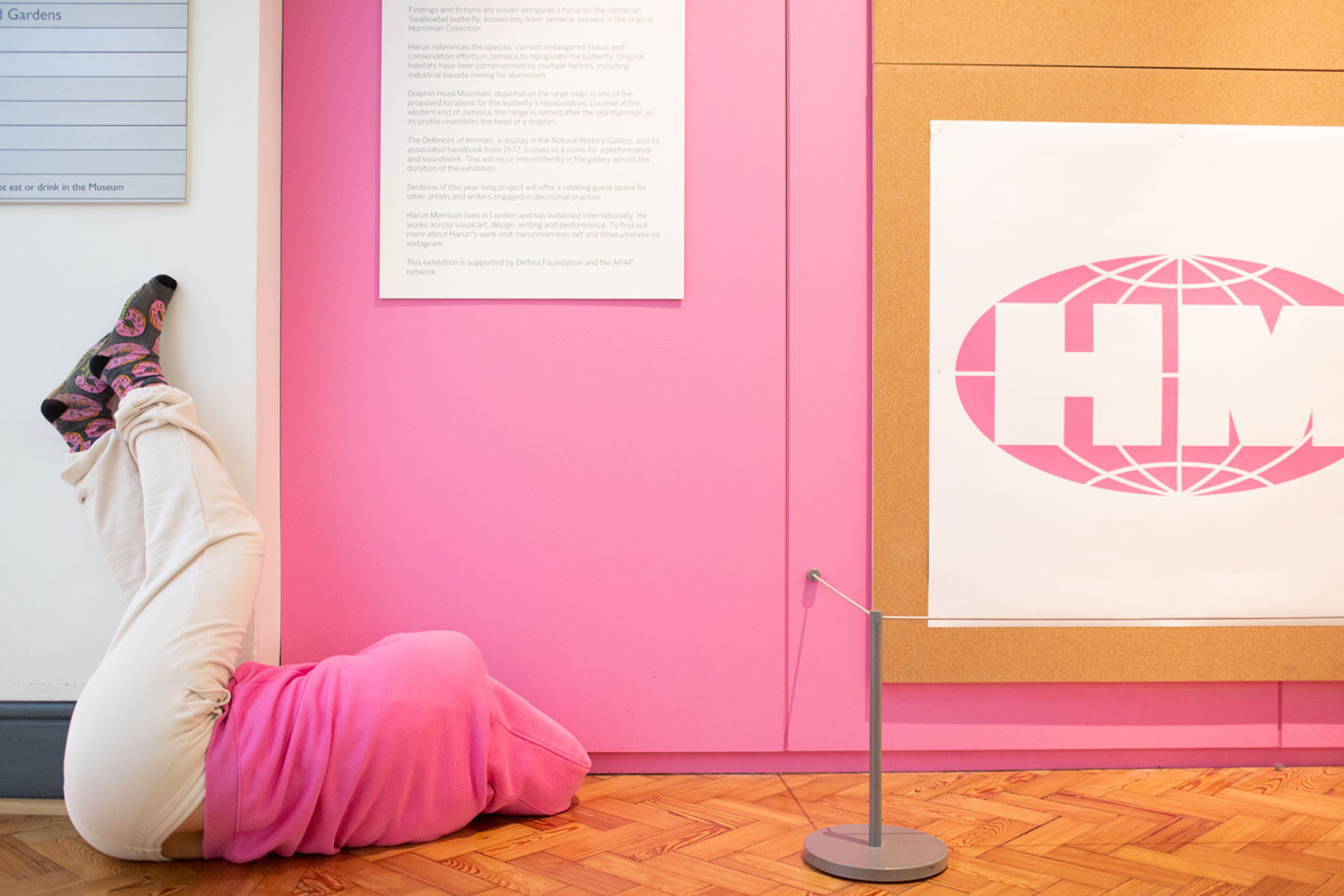 Performer lies on the floor with legs up on a wall. They wear a pink hoodie, which matches the wall.