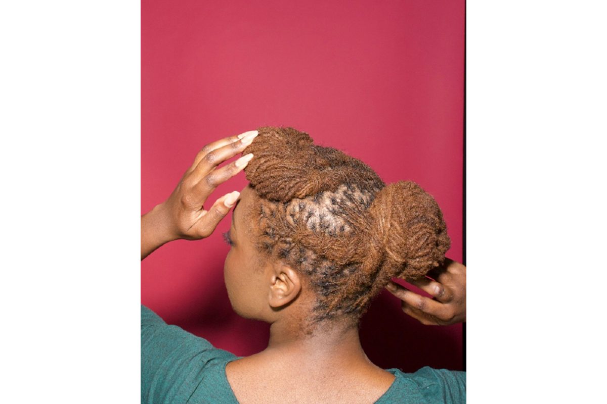 The back of a woman's head with dreadlocks tied into a bun. Her head is turned slightly to the side, and her hand rests on the front of her hair