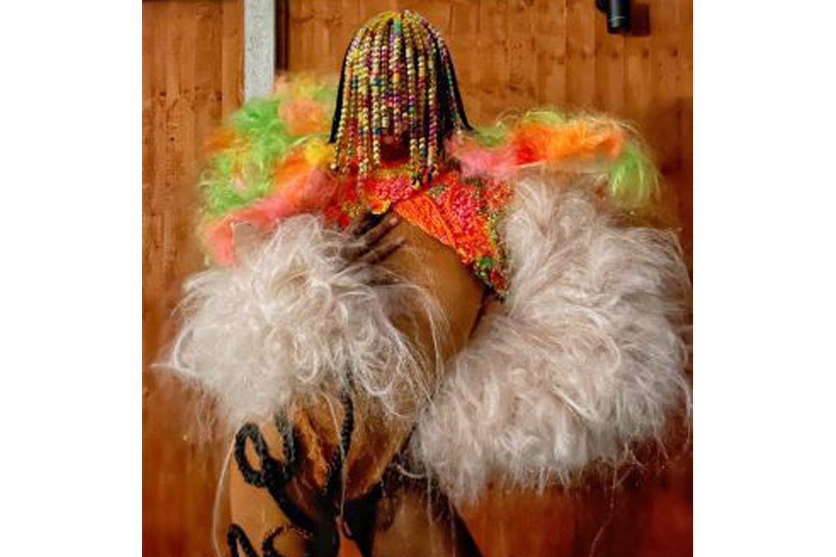 A Black woman wears a colourful feathery top with a white feathery shrug. She has colourful braids which cover her face