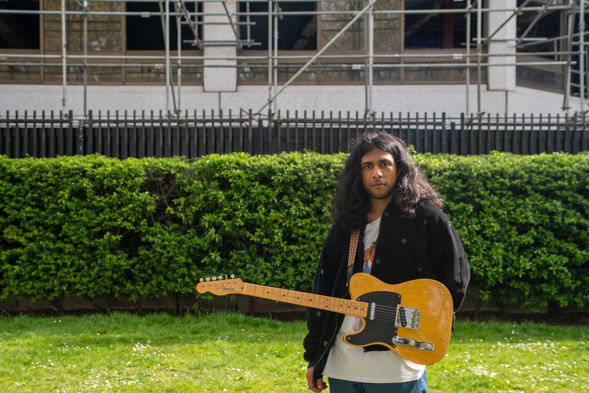 A man with long hair and wearing a black jacket holds a guitar outside