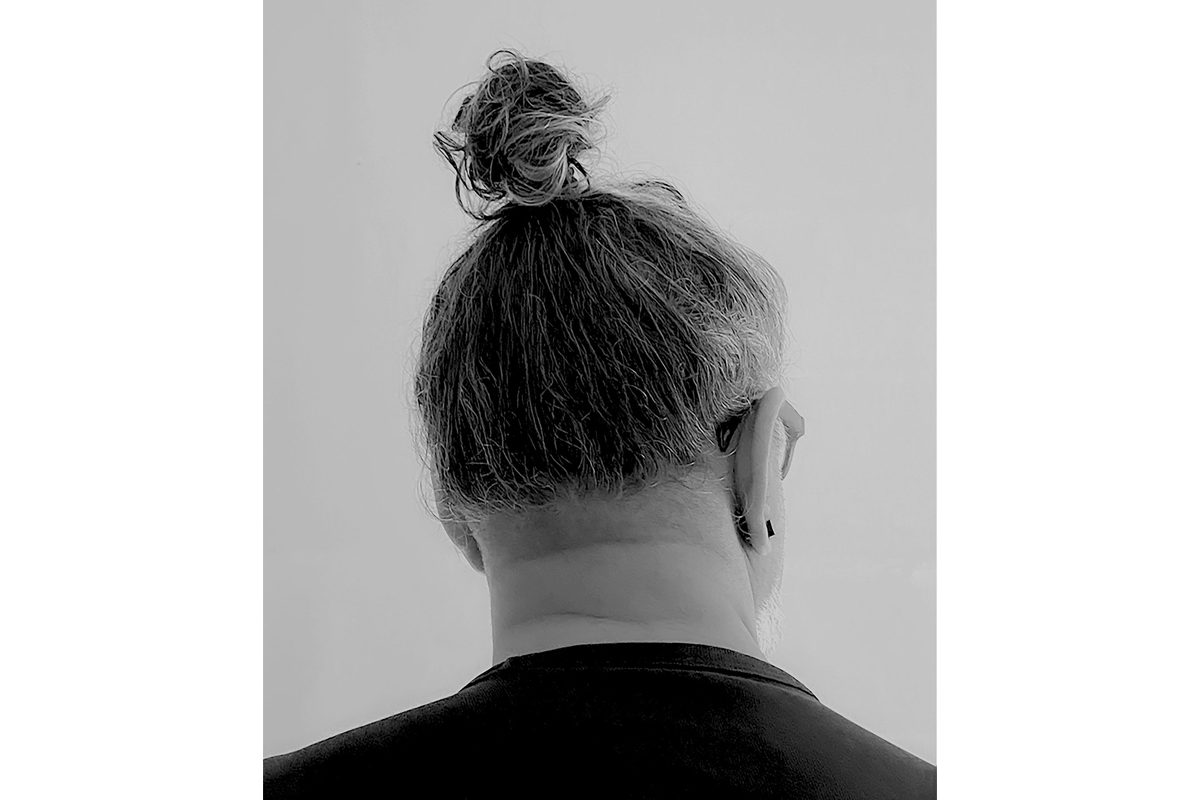 The back of a man's head whose hair is tied in a topknot. He wears a black tshirt and we can see he wears glasses