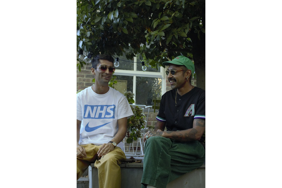 Two men sit next to each other outside, one wears a t-shirt that says 'NHS' with a Nike tick underneath and the other wears a green cap