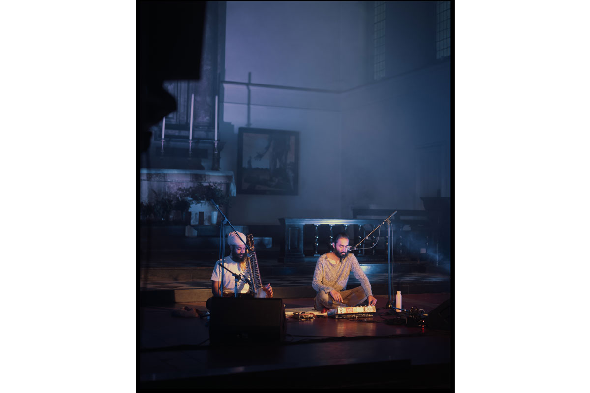 Two men in a large performance space sit on the floor as they play music