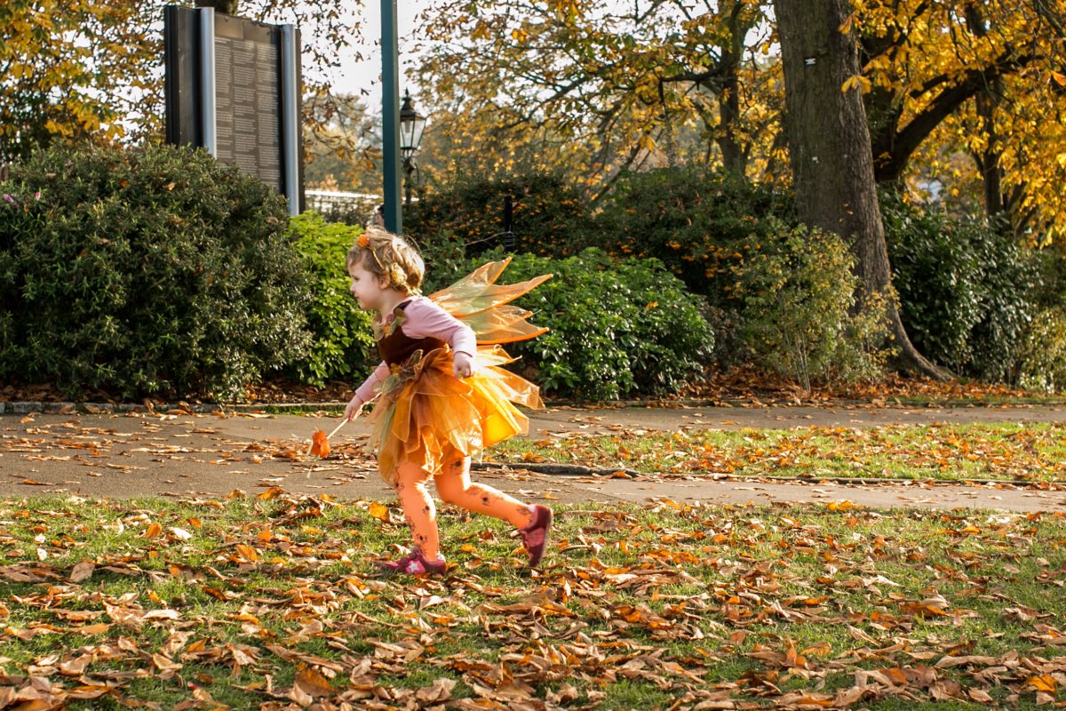 A girl in an orange fairy dress runs through leafy grass at the Horniman
