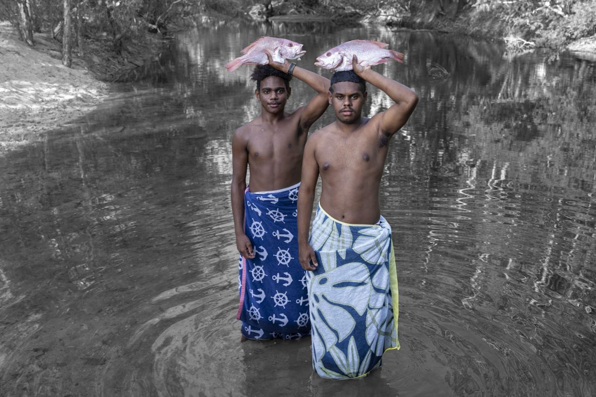 Two boys stand knee deep in a river, holding fish above their heads. They and the fish are in colour but the rest of the image is in black and white.
