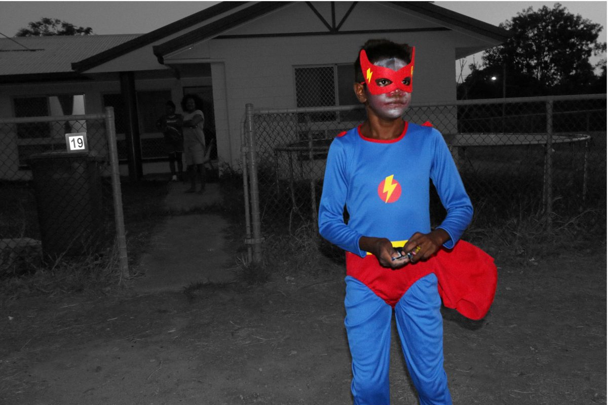 A boy wears a superhero costume including mask in front of a house. He is in colour but the rest of the image is in black and white.