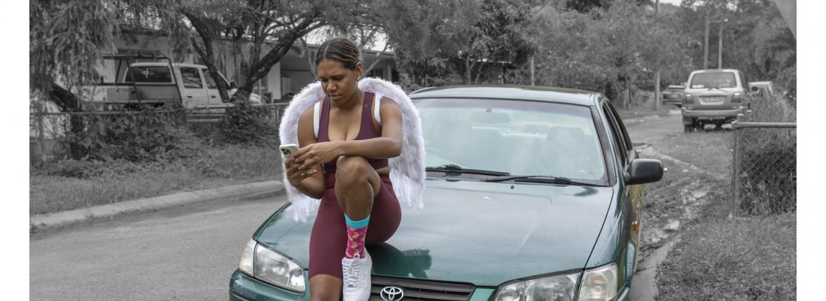 Young girl sits on a car, wearing angel wings and looking at her phone. She and the car are in colour and the rest of the image is in black and white.