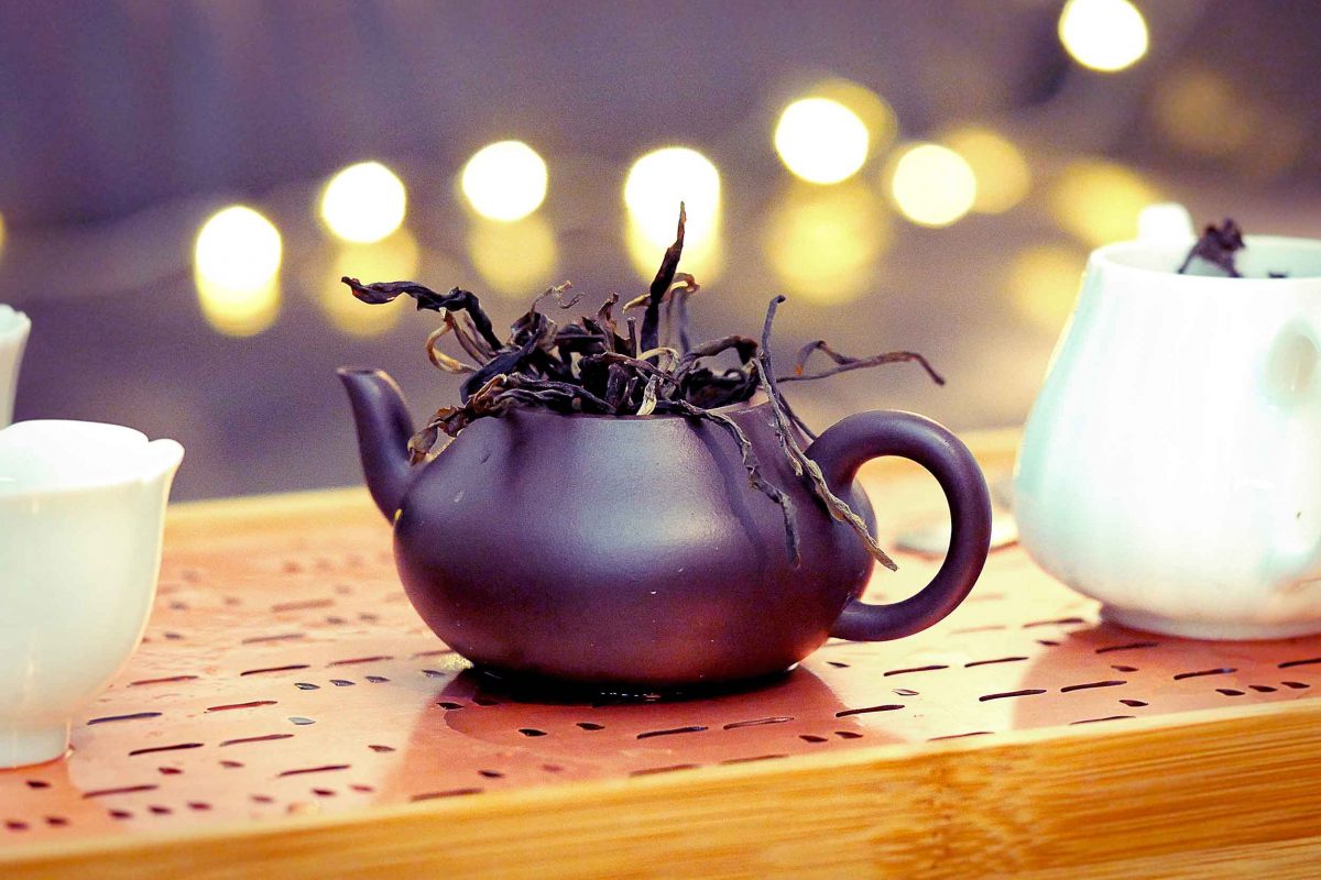 A photo of a teapot with dried tea leaf tendrils spilling from the top. It is sat on a wooden work surface with twinkling lights in the background