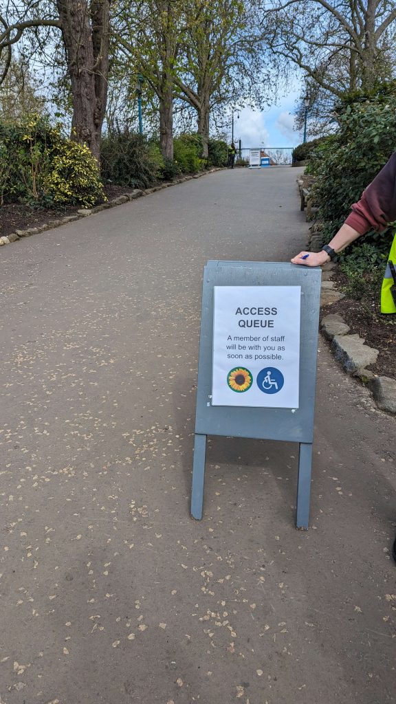 Small board with sign on it reading 'Access Queue a member of staff will be with you as soon as possible' accompanied by a picture of a sunflower and a symbol of a person in a wheelchair