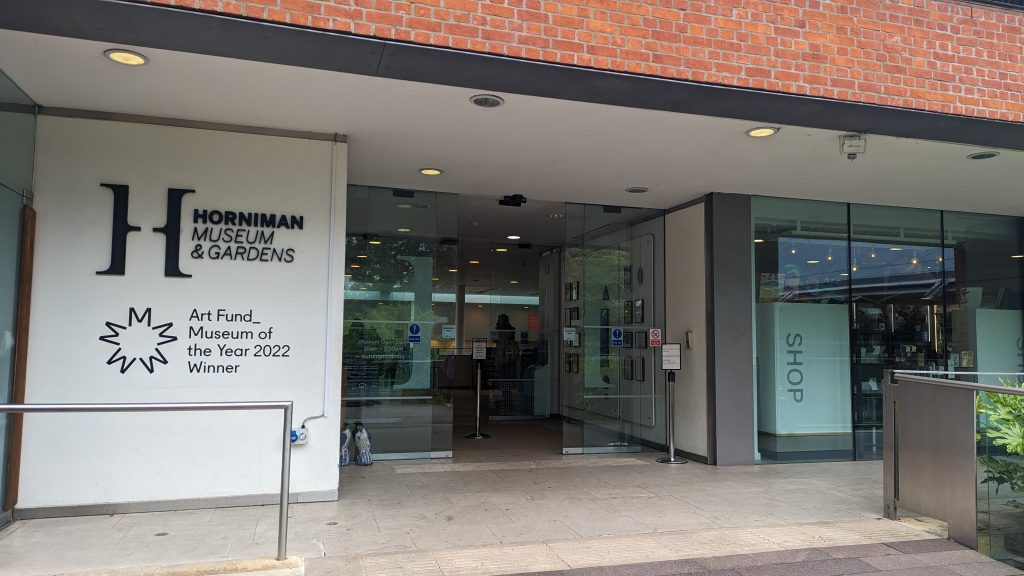 Entrance to the Horniman Museum, with glass entrance doors and a window to the right. To the left of the glass doors is a white wall with text on it which reads 'Horniman Museum and Gardens Art Fund Museum of the Year 2022'