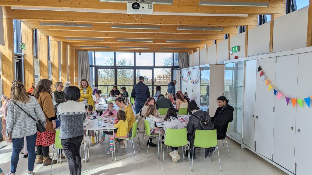 Inside a light room with a big glass window at the back. It is filled with tables and chairs and people sat at them
